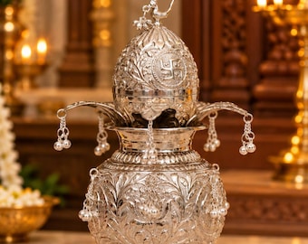 May include: A silver-colored metal kalash vessel featuring intricate floral engravings and a matching lid topped with a bird figure. The vessel is adorned with small hanging silver bells on chains around the rim and the main body, with a swastika symbol embossed on the lid.