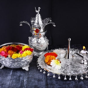 May include: A silver ceremonial set with a decorative pot, a plate with a small bowl, a diya, and a small silver pitcher. The set is decorated with floral patterns and has a traditional Indian design. The set is filled with colorful flowers.