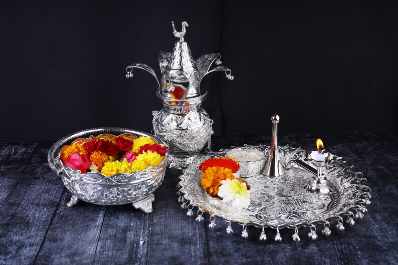 May include: A silver ceremonial set with a decorative pot, a plate with a small bowl, a diya, and a small silver pitcher. The set is decorated with floral patterns and has a traditional Indian design. The set is filled with colorful flowers.