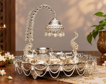 May include: A silver-toned decorative tray featuring an arched peacock-shaped stand with a hanging bell-shaped ornament. The tray includes a small central vessel, a shallow bowl, and is adorned with dangling white beaded fringe along the edges.