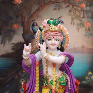 Puede incluir: Una estatua de m&aacute;rmol blanco de Krishna, una deidad hind&uacute;, tocando la flauta. Est&aacute; adornado con ropa colorida, joyas y un tocado de plumas de pavo real. La estatua est&aacute; colocada sobre un fondo pintado de un &aacute;rbol con flores rosas.