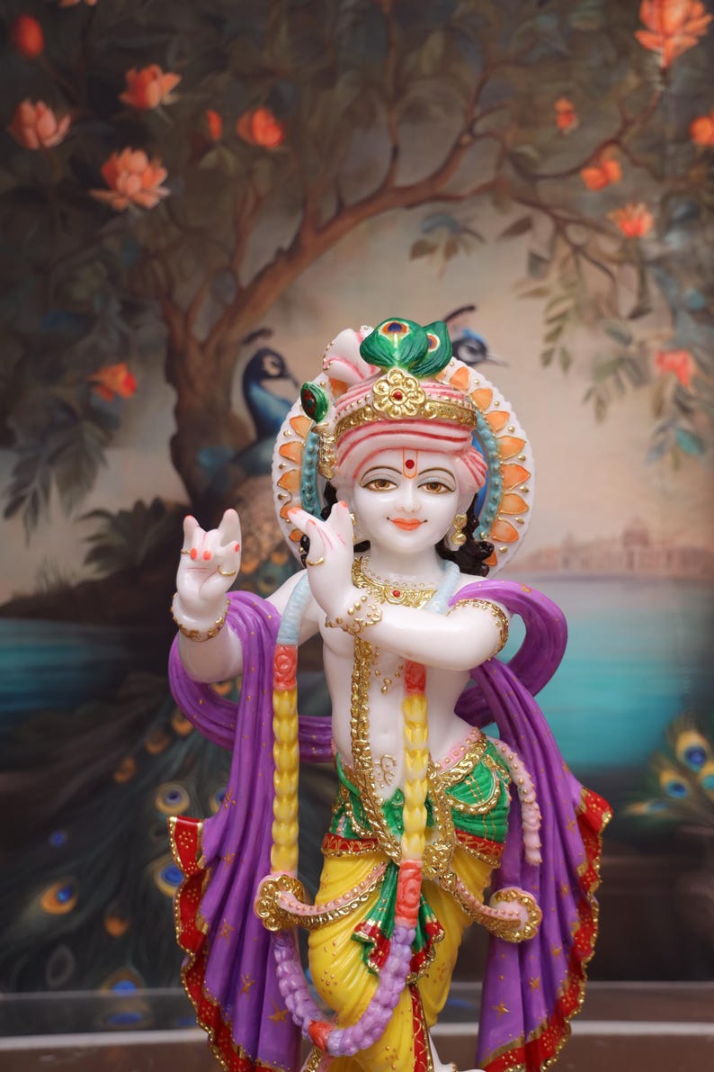 Puede incluir: Una estatua de m&aacute;rmol blanco de Krishna, una deidad hind&uacute;, tocando la flauta. Est&aacute; adornado con ropa colorida, joyas y un tocado de plumas de pavo real. La estatua est&aacute; colocada sobre un fondo pintado de un &aacute;rbol con flores rosas.