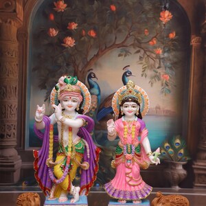 Puede incluir: Un par de estatuas hind&uacute;es de colores, Krishna y Radha, de pie sobre bases blancas con detalles ornamentados. Las estatuas est&aacute;n hechas de m&aacute;rmol blanco y est&aacute;n decoradas con colores vibrantes, incluyendo rosa, amarillo, verde y azul. Dos peque&ntilde;os elefantes de madera se colocan frente a las estatuas.