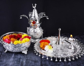 May include: A silver tray with a silver pitcher, a silver bowl filled with yellow and red flowers, and a silver diya with a lit candle. The tray is decorated with intricate designs.