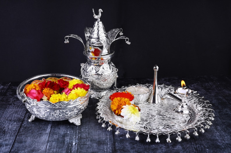 May include: A silver tray with a silver pitcher, a silver bowl filled with yellow and red flowers, and a silver diya with a lit candle. The tray is decorated with intricate designs.