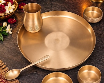 May include: A brass dinnerware set consisting of a large round plate, two small bowls, a drinking cup, and a spoon. Each piece features a smooth, polished gold-toned finish with intricate floral engravings along the lower edges of the bowls and cup.