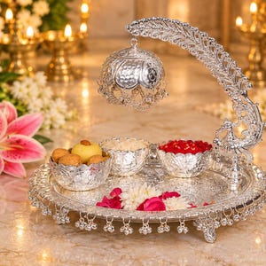 May include: A silver-toned decorative tray featuring an ornate peacock-shaped handle, a hanging bell-shaped dome, and two small bowls. The tray is embellished with dangling metallic charms along the edge and contains small food items and flower petals.