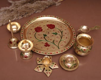 May include: A gold-colored metal puja thali set with a decorative floral design. The set includes a plate, a diya, a bell, a spoon, a kalash, and a small bowl.