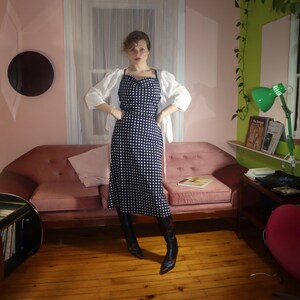 May include: A navy blue and white polka dot dress with spaghetti straps and a white jacket. The dress is paired with black boots. The background includes a pink sofa, wooden floor, and a window.