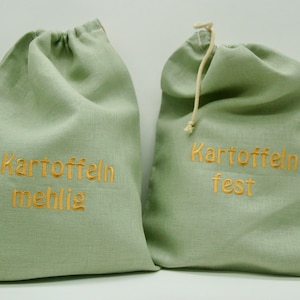 May include: Two sage green drawstring bags with gold embroidered text. One bag reads "Kartoffeln mehlig" and the other reads "Kartoffeln fest". The bags are made of a textured fabric and have a natural-colored drawstring.