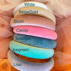 May include: A stack of seven velvet headbands in various colors: white, beige gold, blush, candy, taleblue, icegreen, and lilac. Each headband is labeled with its color name. The headbands are arranged on a peach-colored tulle fabric.