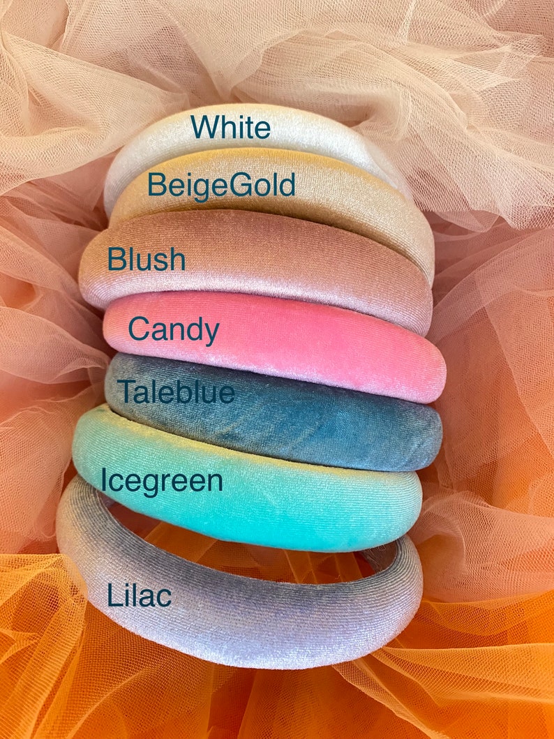 May include: A stack of seven velvet headbands in various colors: white, beige gold, blush, candy, taleblue, icegreen, and lilac. Each headband is labeled with its color name. The headbands are arranged on a peach-colored tulle fabric.