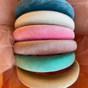 May include: A stack of six velvet headbands in various colors: teal, off-white, pink, rose, mint green, and tan. The headbands are arranged in a neat pile, showcasing their plush texture and vibrant hues. The background is a soft, sheer fabric.