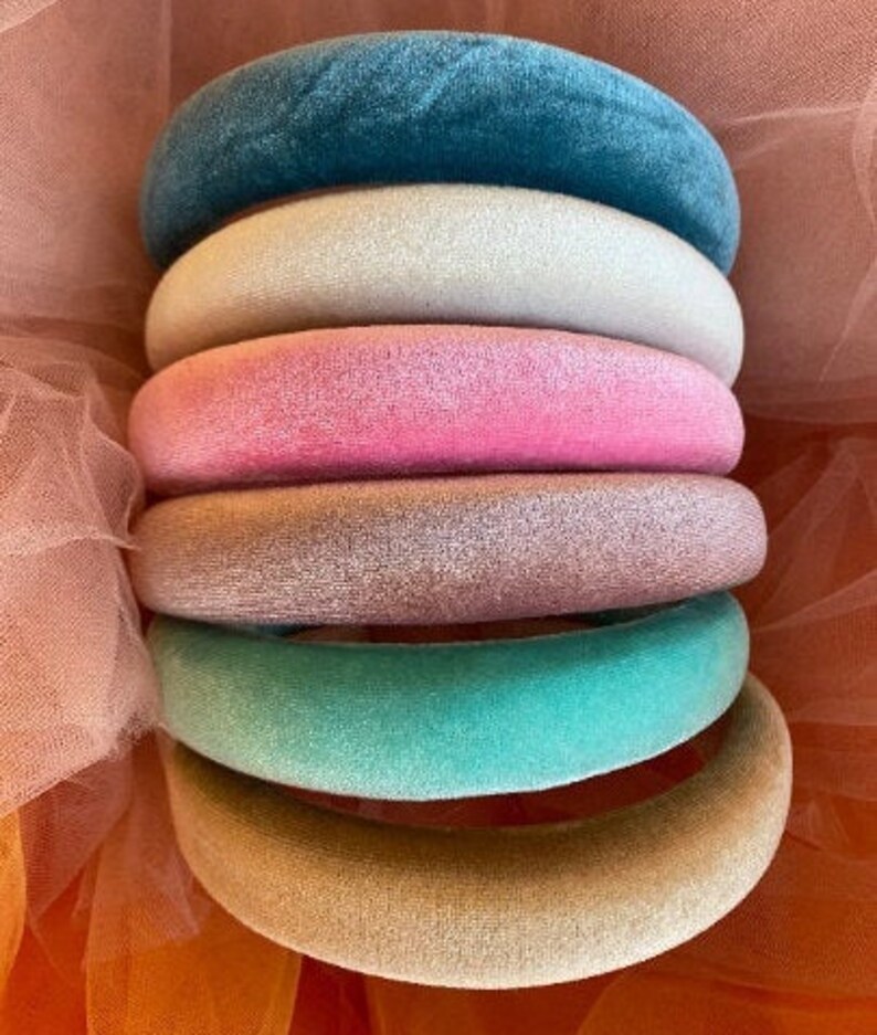 May include: A stack of six velvet headbands in various colors: teal, off-white, pink, rose, mint green, and tan. The headbands are arranged in a neat pile, showcasing their plush texture and vibrant hues. The background is a soft, sheer fabric.