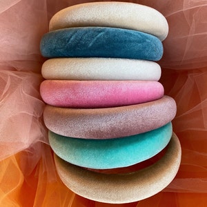 May include: A stack of seven velvet headbands in various colors, including cream, teal, white, pink, mauve, turquoise, and tan. The headbands are arranged in a cascading display, showcasing their plush texture and vibrant hues. The background is a soft orange fabric.