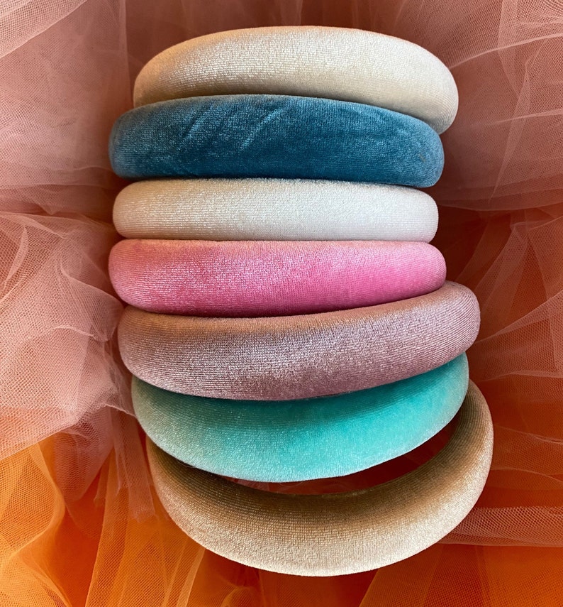 May include: A stack of seven velvet headbands in various colors, including cream, teal, white, pink, mauve, turquoise, and tan. The headbands are arranged in a cascading display, showcasing their plush texture and vibrant hues. The background is a soft orange fabric.