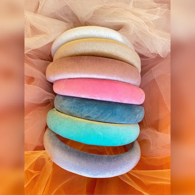 May include: A stack of seven velvet headbands in various colors, including white, yellow, tan, pink, teal, turquoise, and lavender. The headbands are arranged on a backdrop of peach-colored tulle.
