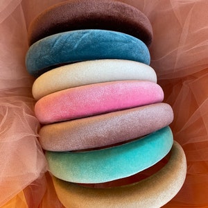 May include: A stack of velvet headbands in various colors, including brown, teal, white, pink, mauve, seafoam green, and beige. The headbands are round and plush, with a soft, velvety texture. The background is a sheer, peach-colored fabric.