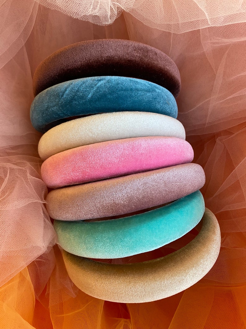 May include: A stack of velvet headbands in various colors, including brown, teal, white, pink, mauve, seafoam green, and beige. The headbands are round and plush, with a soft, velvety texture. The background is a sheer, peach-colored fabric.