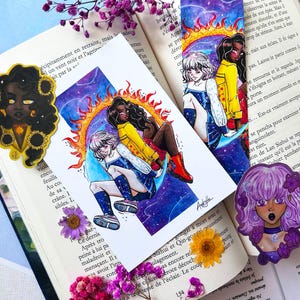 Op de afbeelding: Een collectie kunst in anime-stijl met twee stickers van personages met zonnebloemen en paarse bloemen, een rechthoekige print van twee personages rug aan rug tegen een zon- en sterrenstelselachtergrond, en een boekenlegger met soortgelijke kunst.