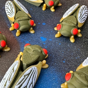 Könnte beinhalten: Fünf grüne und goldene Zikadenfiguren mit weißen Flügeln und roten Augen. Die Zikaden sind in einer Reihe auf einer blau-grünen, gefleckten Oberfläche angeordnet.