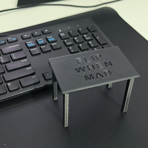 May include: A small, black, rectangular table-shaped object with four legs, featuring the embossed text "FLIP WHEN MAD" on its top surface. The object is constructed from a matte material with visible 3D printed layer lines on the legs and top.