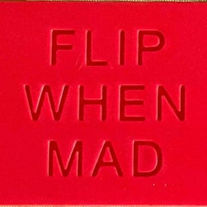 May include: A rectangular red sign featuring the text FLIP WHEN MAD embossed in a bold, sans-serif typeface. The surface is a solid, vibrant red colour, and the lettering is indented into the material, creating a clean and legible visual contrast.