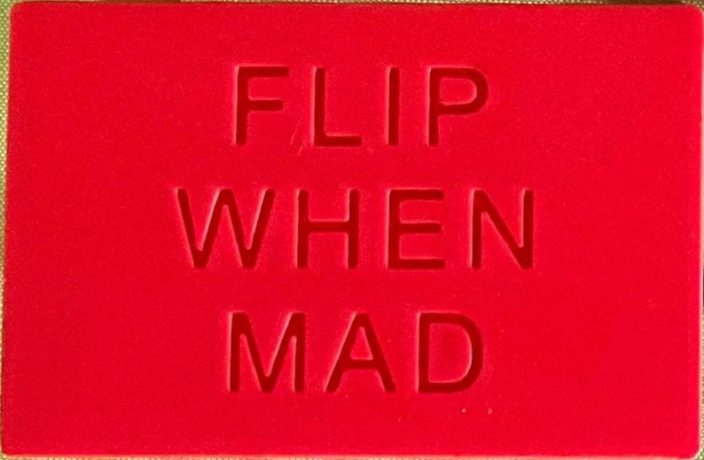 May include: A rectangular red sign featuring the text FLIP WHEN MAD embossed in a bold, sans-serif typeface. The surface is a solid, vibrant red colour, and the lettering is indented into the material, creating a clean and legible visual contrast.