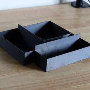 May include: A black desk organizer with multiple compartments. The organizer is made of a matte black material and has a rectangular shape with dividers to separate items. It sits on a light wood surface.