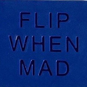 May include: A rectangular blue sign with the text FLIP WHEN MAD embossed in a bold, sans-serif font. The surface has a uniform matte finish, and the letters are recessed into the material, creating a clean, high-contrast visual appearance.