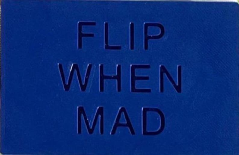 May include: A rectangular blue sign with the text FLIP WHEN MAD embossed in a bold, sans-serif font. The surface has a uniform matte finish, and the letters are recessed into the material, creating a clean, high-contrast visual appearance.