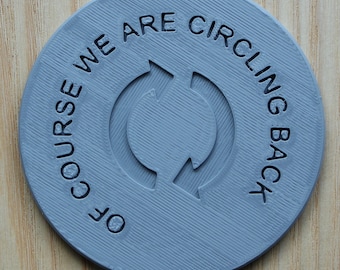 May include: A circular light blue coaster with a textured surface featuring a central icon of two curved arrows forming a cycle. The text "OF COURSE WE ARE CIRCLING BACK" is embossed in a circular pattern around the central arrow icon in dark blue.