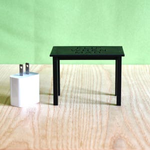 May include: A small black rectangular table with four legs and the text "FLIP WHEN MAD" engraved on the top surface, positioned next to a white rectangular power adapter with two metal prongs extending from the top.