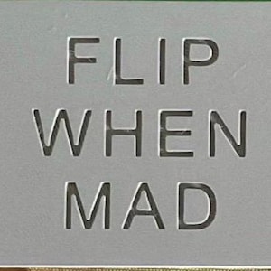 May include: A rectangular grey sign with the text FLIP WHEN MAD engraved in a clean, sans-serif font. The surface is smooth and matte, featuring dark, recessed lettering that stands out against the light grey background of the flat object.