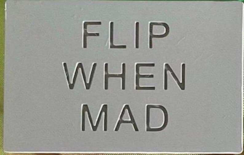 May include: A rectangular grey sign with the text FLIP WHEN MAD engraved in a clean, sans-serif font. The surface is smooth and matte, featuring dark, recessed lettering that stands out against the light grey background of the flat object.