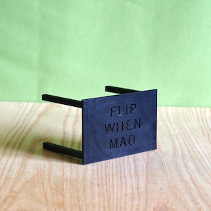 May include: A black rectangular stand with a flat front panel featuring the embossed text "FLIP WHEN MAD" in a sans-serif font. The object has two horizontal support bars extending backward from the sides of the main vertical panel.