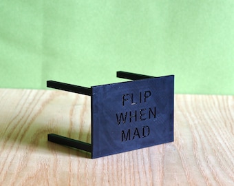 May include: A black rectangular stand with a flat front panel featuring the embossed text "FLIP WHEN MAD" in a sans-serif font. The object has two horizontal support bars extending backward from the sides of the main vertical panel.