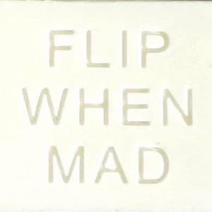 May include: A rectangular off-white sign featuring the text FLIP WHEN MAD in embossed, light-coloured capital letters. The surface has a subtle texture, and the lettering is centred, creating a clean, monochromatic appearance across the entire piece.