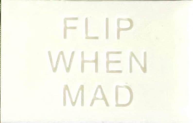 May include: A rectangular off-white sign featuring the text FLIP WHEN MAD in embossed, light-coloured capital letters. The surface has a subtle texture, and the lettering is centred, creating a clean, monochromatic appearance across the entire piece.