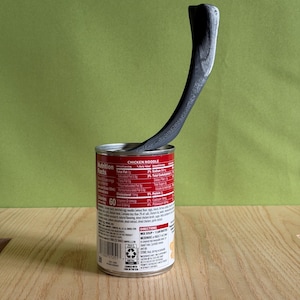 May include: A black, curved, textured object resembling a wishbone is inserted into a red and white metal soup can labeled Chicken Noodle. The can features a nutrition facts label with text including Total Fat 2g, Sodium 890mg, and Calories 60.