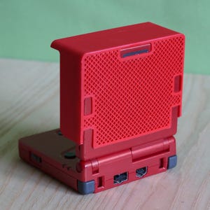 May include: A red handheld gaming console with a hinged design, featuring a bright red protective cover with a textured grid pattern attached to the top lid. The base of the device shows grey hinge accents and ports along the back edge.
