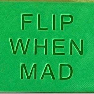 May include: A rectangular green object with the text FLIP WHEN MAD embossed in a sans-serif font on the front surface. The lettering is recessed into the material, creating a clean and uniform appearance across the entire flat, solid green face.