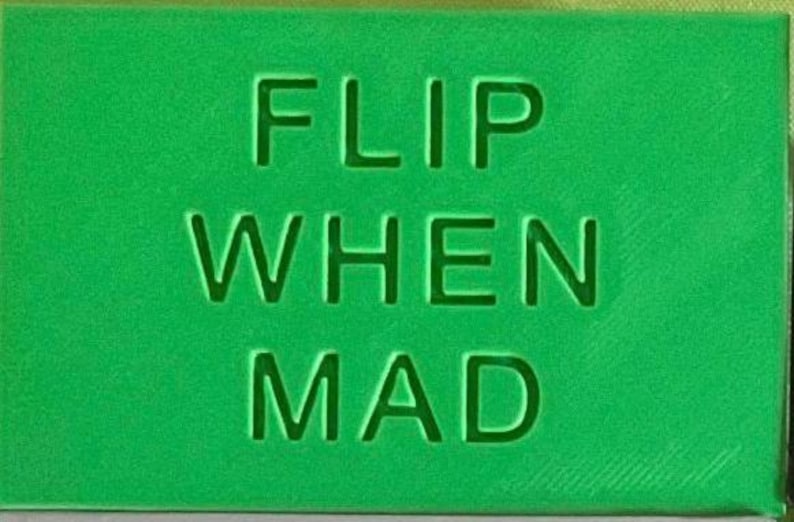 May include: A rectangular green object with the text FLIP WHEN MAD embossed in a sans-serif font on the front surface. The lettering is recessed into the material, creating a clean and uniform appearance across the entire flat, solid green face.