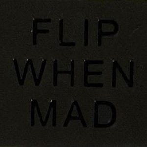 May include: A rectangular black patch featuring the embossed text FLIP WHEN MAD in a bold, sans-serif font. The surface of the patch has a subtle, fine-grained texture, and the lettering is raised, creating a monochromatic and tactile appearance.