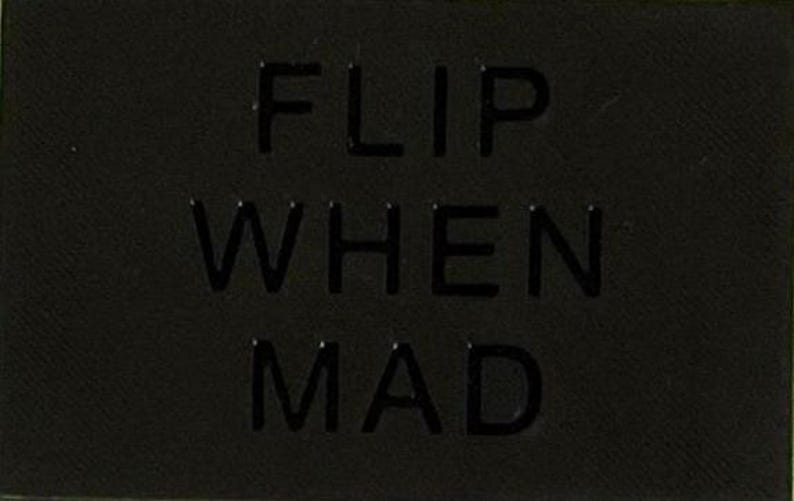May include: A rectangular black patch featuring the embossed text FLIP WHEN MAD in a bold, sans-serif font. The surface of the patch has a subtle, fine-grained texture, and the lettering is raised, creating a monochromatic and tactile appearance.