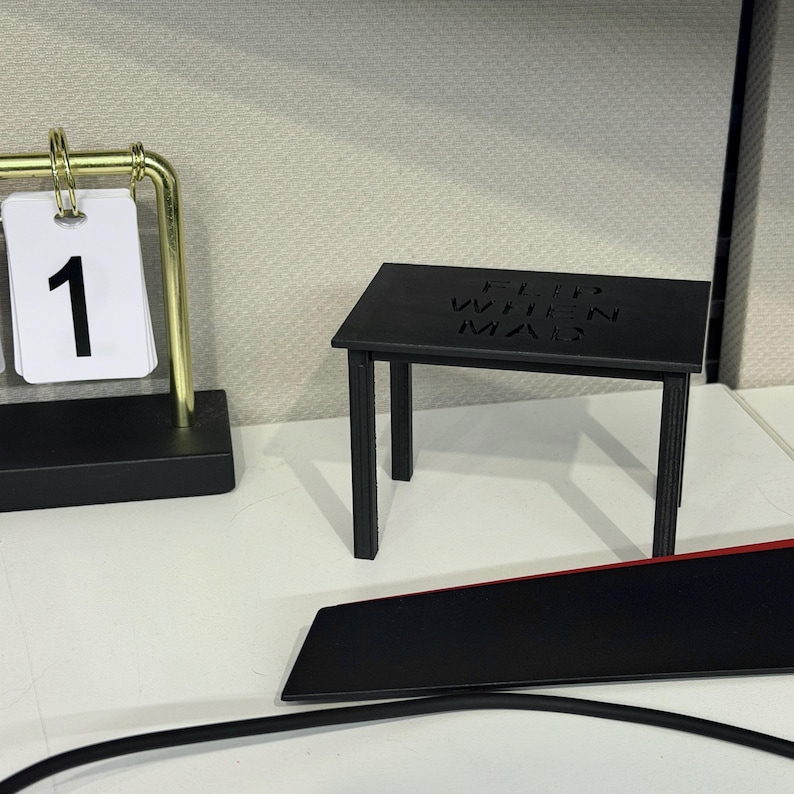 May include: A small black rectangular table with the text FLIP WHEN MAD embossed on the top surface. The table has four thin legs and a flat tabletop, positioned next to a gold-toned metal desk calendar displaying the number one on a white card.