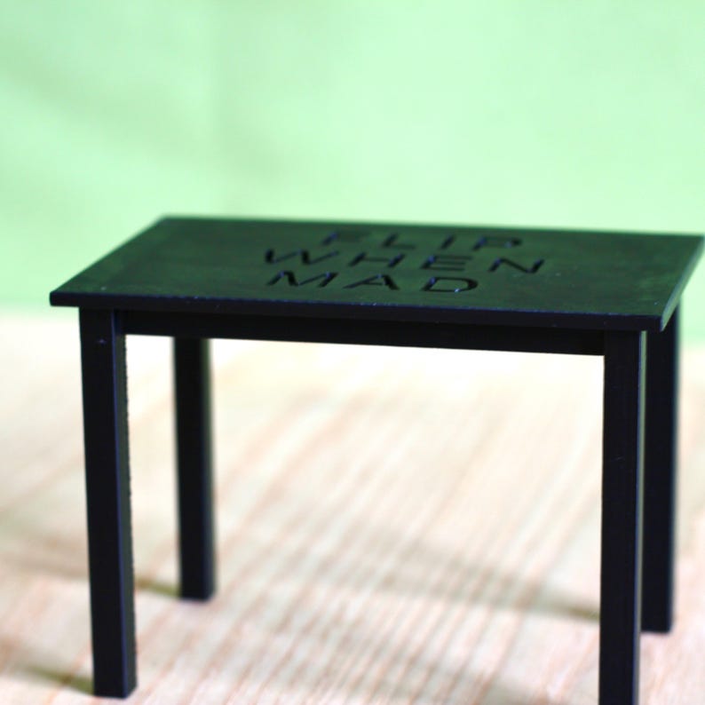 May include: A small black rectangular table with four straight legs. The top surface of the table features the embossed text "FLIP WHEN MAD" centred in a clear, sans-serif font. The object has a matte finish and a simple, functional structure.