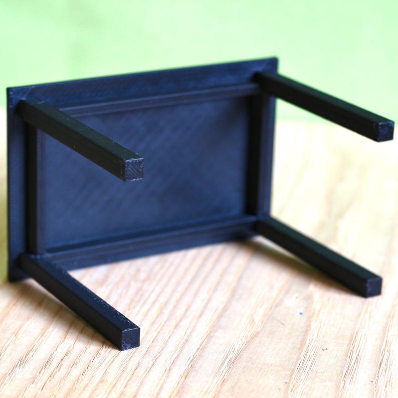 May include: A small, black, rectangular table shown from an inverted perspective. The piece features a flat, solid top surface and four straight, square-profile legs extending upwards, showcasing a uniform matte finish and a simple, clean construction.