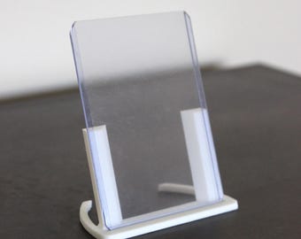 May include: A clear acrylic display stand with a white base and supports. The stand is designed to hold a rectangular item, such as a sign or a photo. The stand is made of plastic and is suitable for displaying items.