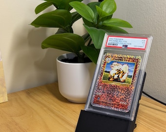 May include: A graded Pokémon card of Arcanine from the Legendary Collection set, displayed in a clear protective case. The card is held by a black stand on a wooden surface, with a potted green plant in the background.